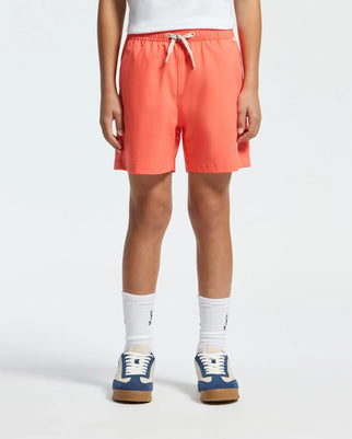 Someone stands before a plain light background wearing vibrant coral Psycho Bunny KIDS SAMOS HYDROCHROMIC PRINTED SWIM TRUNK - B0W453F200, which boasts quick-dry fabric and features a white drawstring. Their look is completed with white socks and blue and white sneakers, while their torso is not visible.