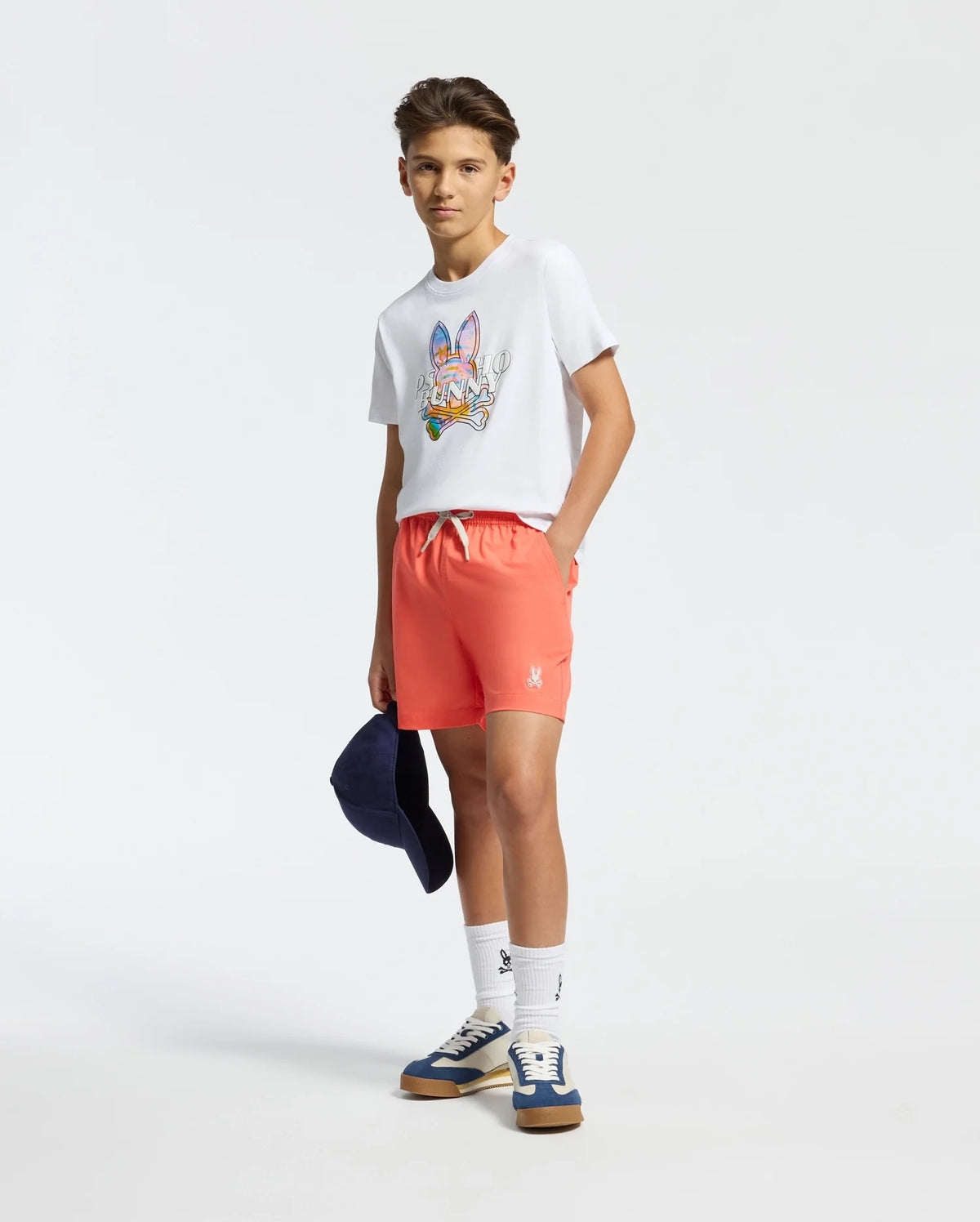 A teen boy, dressed in a casual and sporty outfit, wears a white T-shirt with a colorful graphic and Psycho Bunny's Kids Samos Hydrochromic Printed Swim Trunk in coral, paired with sneakers. He's holding a navy cap against a light background that highlights his clothing.