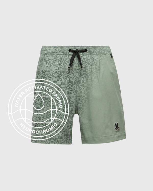 The KIDS SAMOS HYDROCHROMIC PRINTED SWIM TRUNK by Psycho Bunny features a light green hydrochromic fabric with a subtle Bunny pattern, black drawstring, small bunny logo by the hem, and overlay text: “Water Activated Fabric Hydrochromic.” Plain grey background.