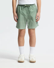 A child wears Psycho Bunny KIDS BENDIGO ALL OVER PRINTED SWIM TRUNK - B0W619F200, paired with white crew socks and white sneakers, standing against a plain light background. Only the lower torso and legs are visible.
