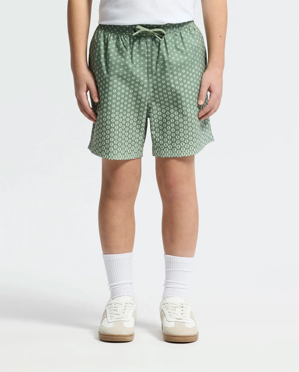 A child wears Psycho Bunny KIDS BENDIGO ALL OVER PRINTED SWIM TRUNK - B0W619F200, paired with white crew socks and white sneakers, standing against a plain light background. Only the lower torso and legs are visible.