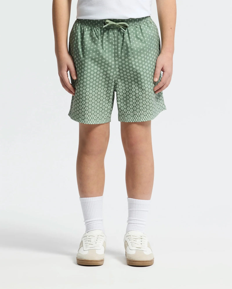 A child wears Psycho Bunny KIDS BENDIGO ALL OVER PRINTED SWIM TRUNK - B0W619F200, paired with white crew socks and white sneakers, standing against a plain light background. Only the lower torso and legs are visible.