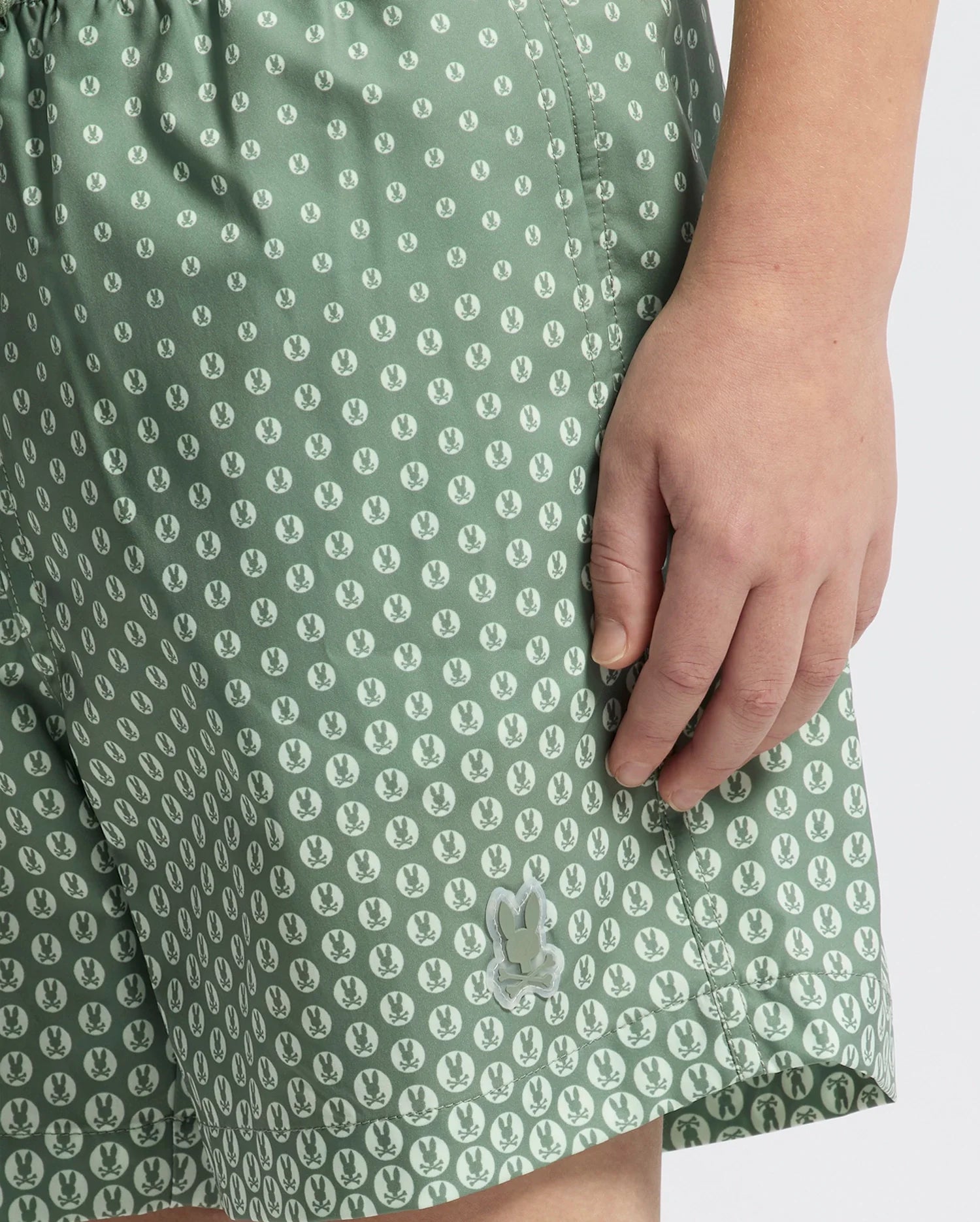 A close-up shows a child wearing Psycho Bunny KIDS BENDIGO ALL OVER PRINTED SWIM TRUNK—green with white bunny and leaf print. Their right hand rests near the pocket, highlighting the quick-dry fabric perfect for active days.