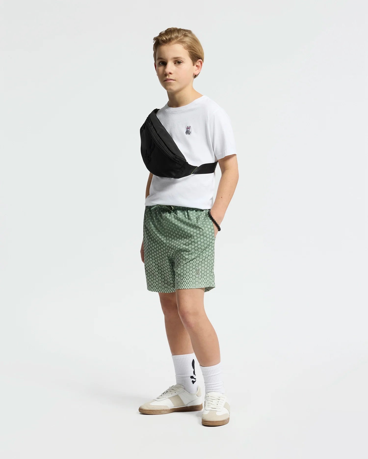 A boy stands against a white background wearing a white t-shirt, Psycho Bunny KIDS BENDIGO ALL OVER PRINTED SWIM TRUNK in green, white sneakers, patterned socks, and a black crossbody bag, looking to the side with his hands in his pockets.