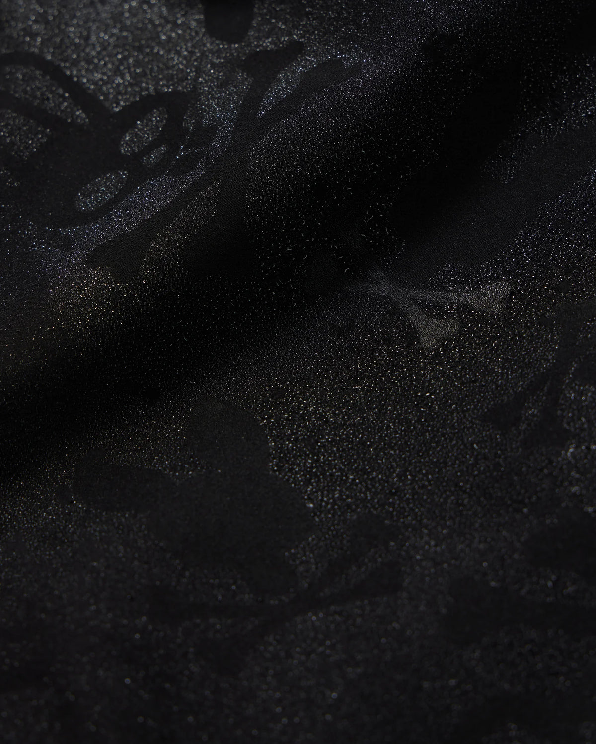 Close-up of Psycho Bunny's KIDS PARKER HYDROCHROMIC SWIM TRUNK (B0W646C200) featuring a subtle, intricate pattern of skulls, bones, and abstract shapes that creates a textured, shadowy effect on the black fabric. The quick-dry material boasts a glossy finish with a slightly shimmering surface.