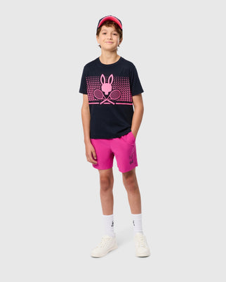 A young person stands confidently against a plain background, wearing a black t-shirt with a pink graphic of a rabbit and crossed tennis rackets. They are also sporting the bright pink KIDS PARKER HYDROCHROMIC SWIM TRUNK by Psycho Bunny, a pink visor, white socks, and white sneakers, with hands in their pockets.