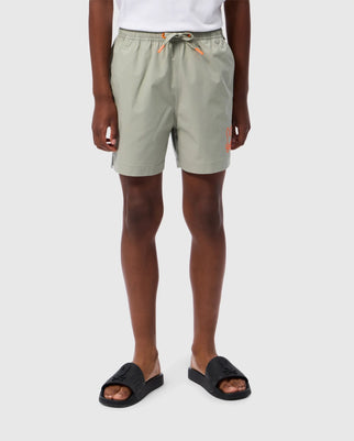 A person is standing against a plain background wearing light green, quick-drying Psycho Bunny KIDS PARKER HYDROCHROMIC SWIM TRUNK - B0W646C200 with orange details and black slide sandals. The photo is cropped at the torso, highlighting visible legs.