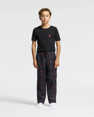 A young boy stands against a plain white background wearing the KIDS TELRIDE PRINTED PAJAMA SET - B0X355E200 by Psycho Bunny. His outfit consists of a black T-shirt with a subtle red logo on the chest, perfectly paired with black pajama pants featuring an eye-catching red pattern. Made from 100% cotton sleepwear, he completes the cozy look with white socks and relaxed hands by his sides.