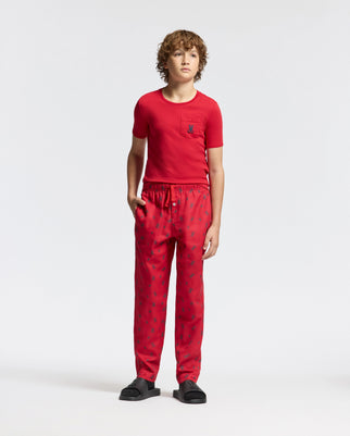 A young person stands against a plain background, wearing the Kids Telride Printed Pajama Set by Psycho Bunny, featuring red t-shirt and pants with subtle patterns. They have one hand in their pocket and are wearing black open-toed sandals.