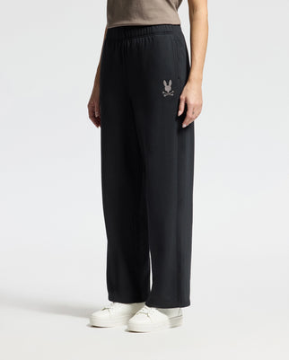 A woman models the Psycho Bunny WOMENS ALEXANDER SWEATPANT - B2P157E200 in a premium cotton blend, standing against a simple background. The sleek black sweatpants display a subtle embroidered logo on the left thigh and are stylishly paired with white sneakers.