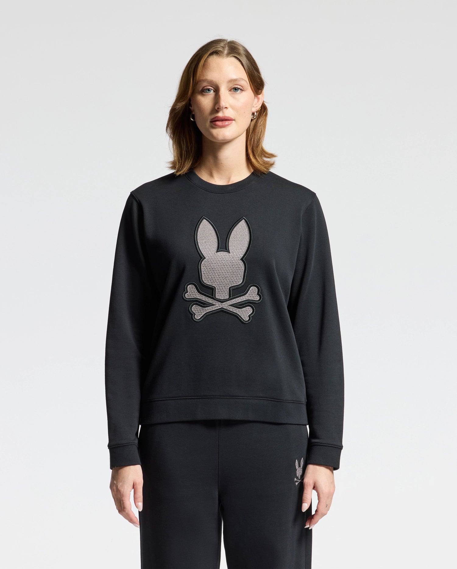A person with shoulder-length hair is wearing a WOMENS ALEXANDER SWEATSHIRT - B2S156E200 from Psycho Bunny, paired with matching pants. The black cotton blend sweatshirt features an embroidered lurex bunny head above crossbones against a plain white background.