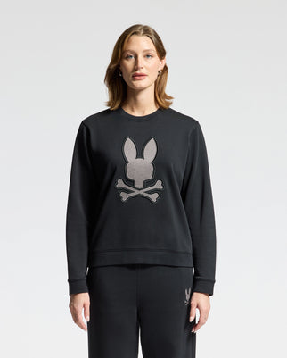 A person with shoulder-length hair is wearing a WOMENS ALEXANDER SWEATSHIRT - B2S156E200 from Psycho Bunny, paired with matching pants. The black cotton blend sweatshirt features an embroidered lurex bunny head above crossbones against a plain white background.