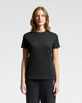 A person with shoulder-length hair is wearing the Psycho Bunny black women's Alexander graphic tee, paired with pants, featuring a small emblem of a bunny with Lurex embroidery on the chest. They stand facing the camera against a plain, light-colored background.