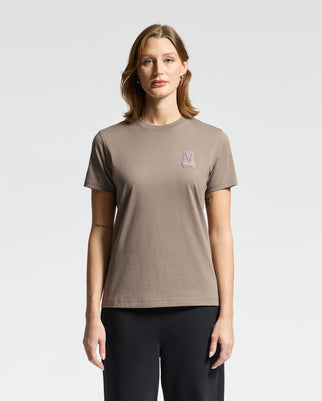 A person wearing the Psycho Bunny WOMENS ALEXANDER GRAPHIC TEE - B2U164E200 in taupe, featuring a small bunny illustration on the chest, stands against a plain white background. The Pima cotton T-shirt is paired with black pants, creating a minimalist yet stylish look.