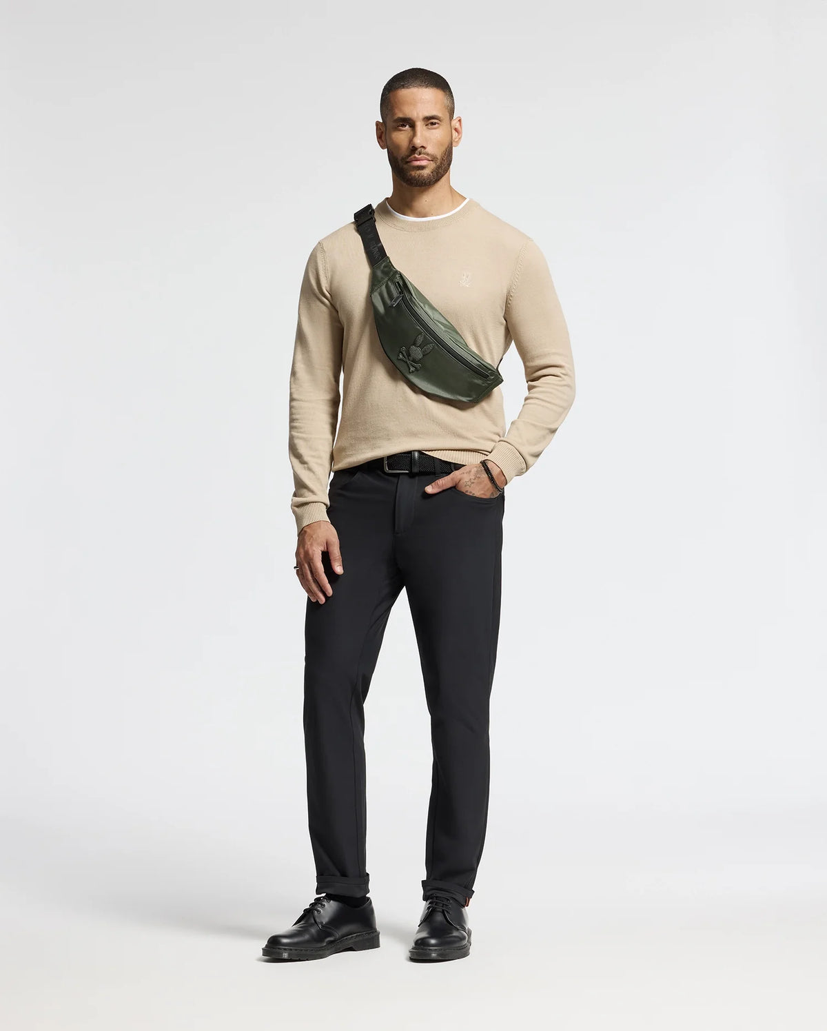 A man stands against a plain background wearing a beige sweater, black pants, and black shoes. He sports the Psycho Bunny MENS CROSS BODY BAG - B6A181E200, crafted from durable nylon, slung over his shoulder.