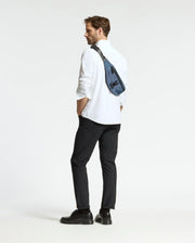 A man stands slightly turned away with hands in pockets, wearing black shoes and a blue Psycho Bunny MENS CROSS BODY BAG - B6A181E200. His white shirt and black pants contrast the plain white background, highlighting his casual look.