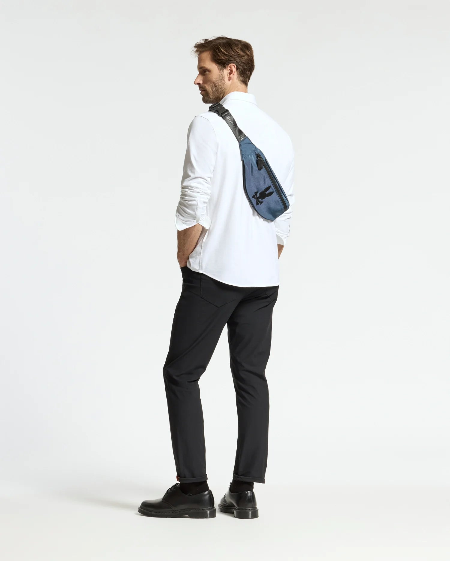 A man stands slightly turned away with hands in pockets, wearing black shoes and a blue Psycho Bunny MENS CROSS BODY BAG - B6A181E200. His white shirt and black pants contrast the plain white background, highlighting his casual look.