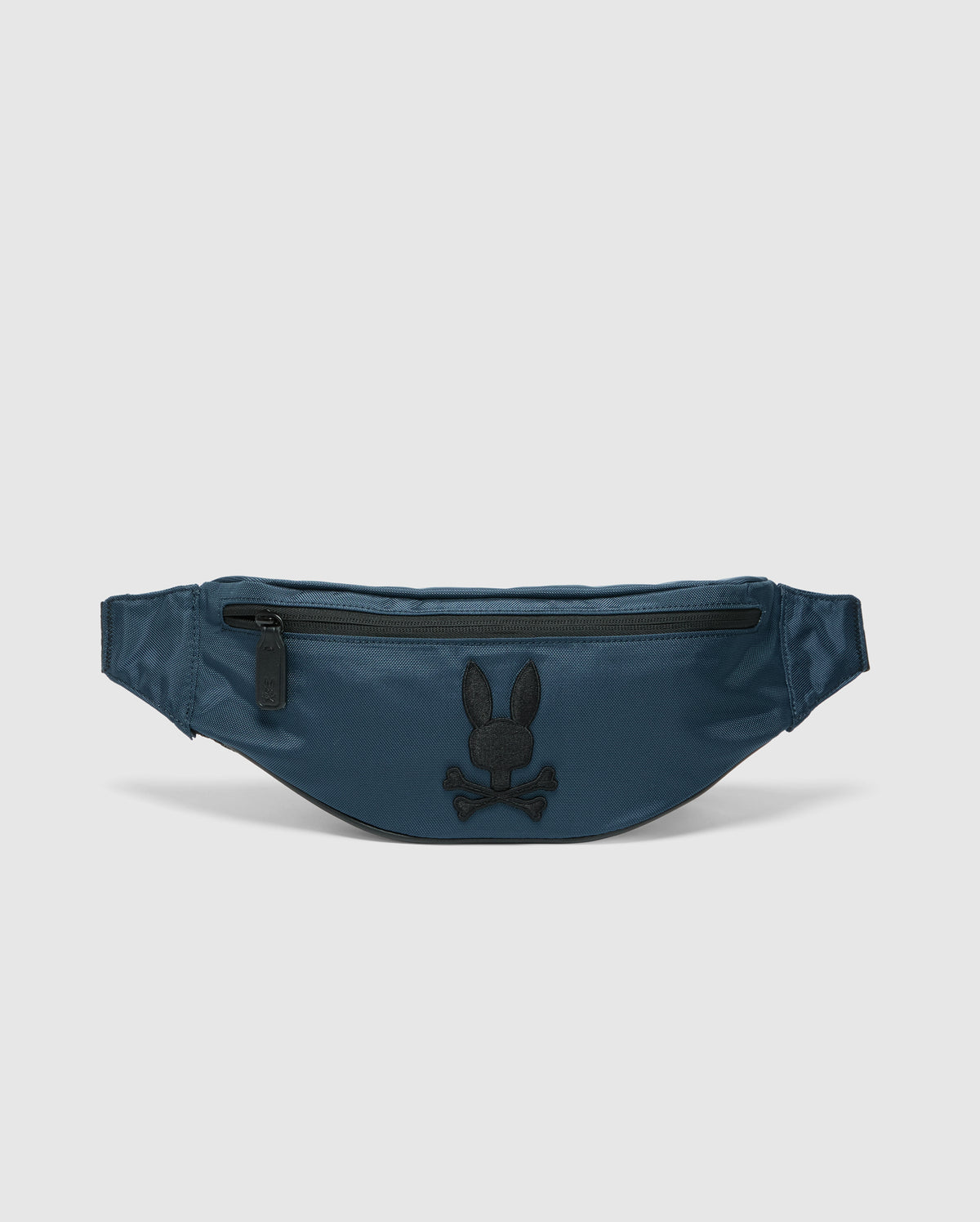 The Psycho Bunny MENS CROSS BODY BAG - B6A181E200, in dark teal nylon, doubles as a fanny pack with a front zipper pocket. It features a black embroidered skull with bunny ears logo and is displayed on a light gray background.