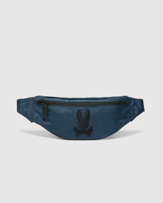 The Psycho Bunny MENS CROSS BODY BAG - B6A181E200, in dark teal nylon, doubles as a fanny pack with a front zipper pocket. It features a black embroidered skull with bunny ears logo and is displayed on a light gray background.