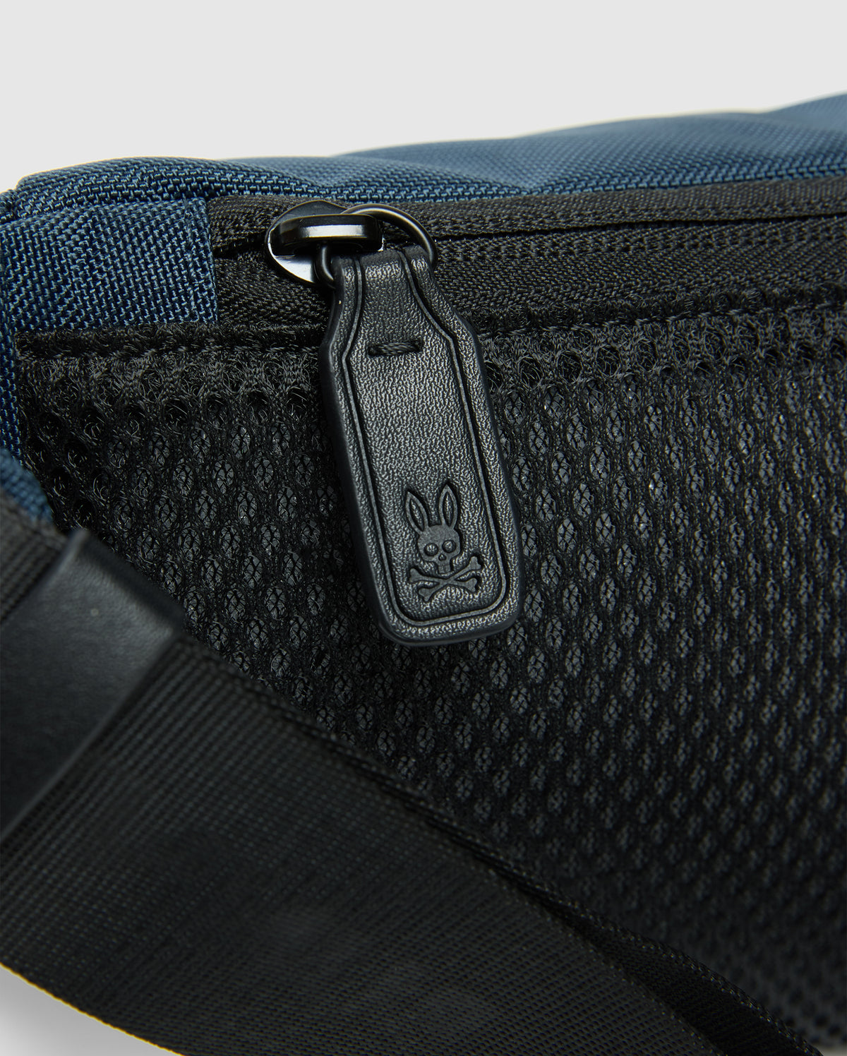 A close-up reveals the Psycho Bunny MENS CROSS BODY BAG - B6A181E200: a blue and black casual bag with a mesh pocket and zipper. The zipper pull is etched with a cartoon rabbit face. A black strap on the lower left enhances its practical nylon design.