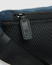 A close-up reveals the Psycho Bunny MENS CROSS BODY BAG - B6A181E200: a blue and black casual bag with a mesh pocket and zipper. The zipper pull is etched with a cartoon rabbit face. A black strap on the lower left enhances its practical nylon design.