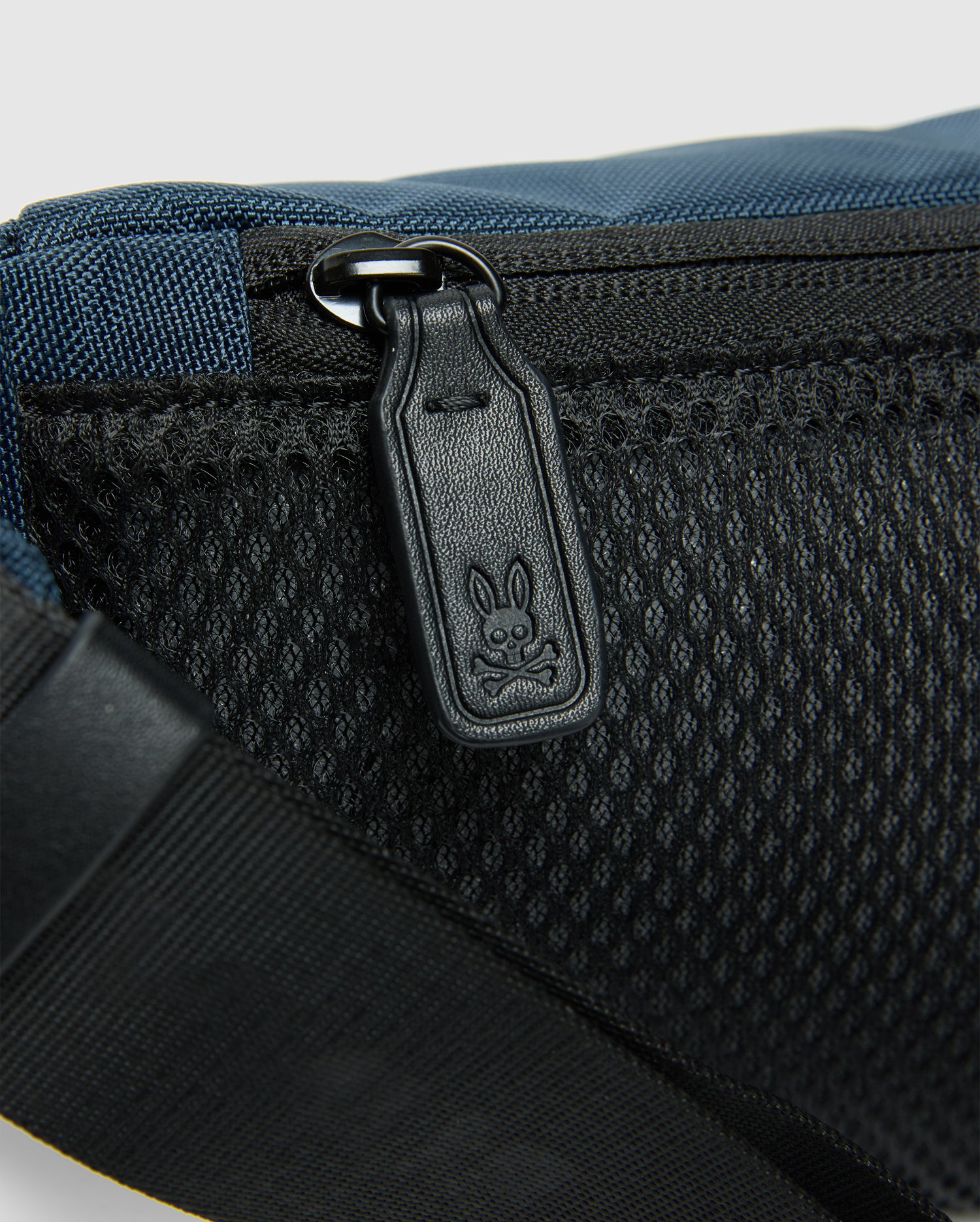 A close-up reveals the Psycho Bunny MENS CROSS BODY BAG - B6A181E200: a blue and black casual bag with a mesh pocket and zipper. The zipper pull is etched with a cartoon rabbit face. A black strap on the lower left enhances its practical nylon design.