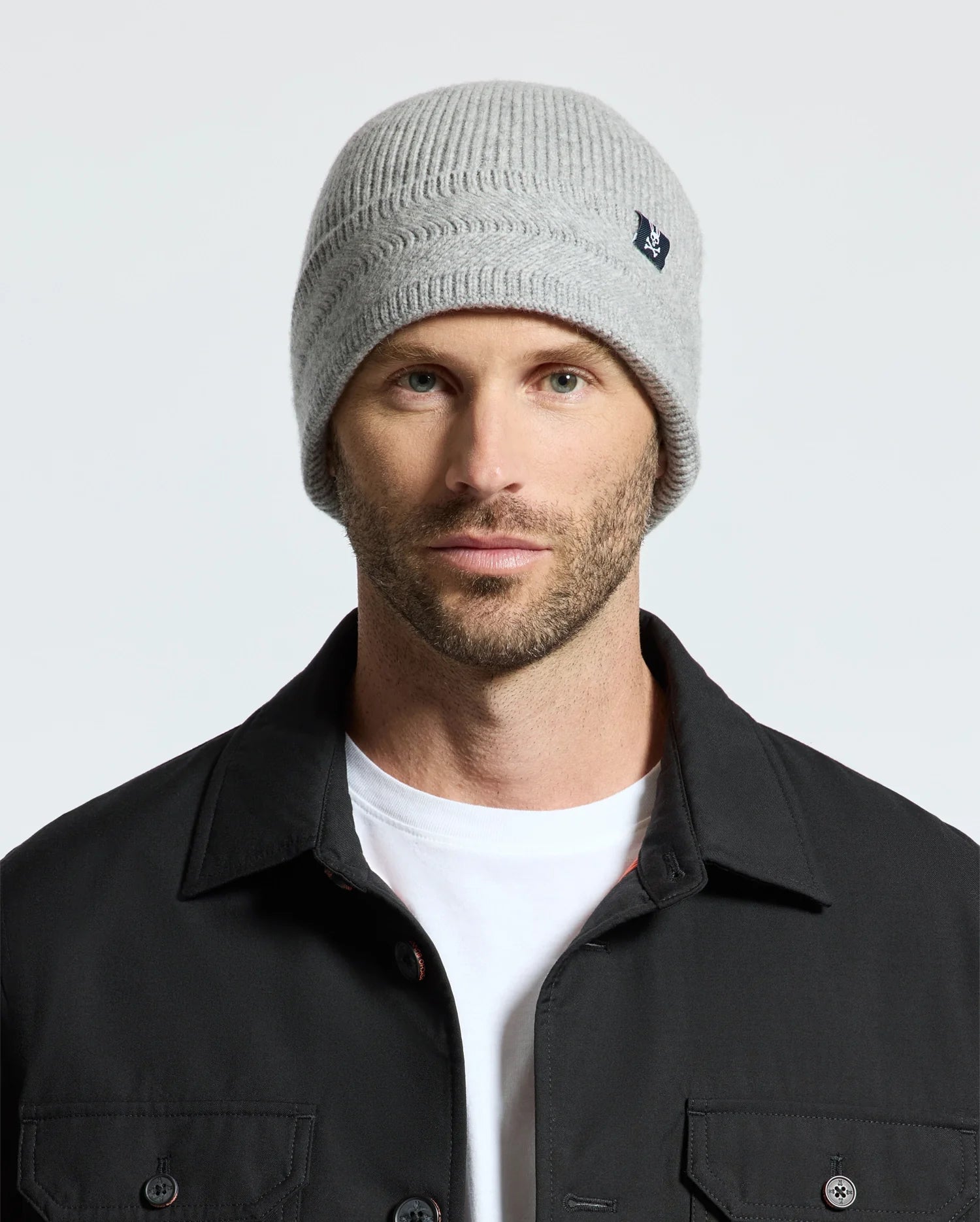 A man wearing a light gray Psycho Bunny MENS RACKED BEANIE - B6A188E200, a black jacket, and a white t-shirt. The background is plain and light-colored.