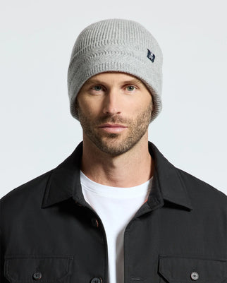 A man wearing a light gray Psycho Bunny MENS RACKED BEANIE - B6A188E200, a black jacket, and a white t-shirt. The background is plain and light-colored.