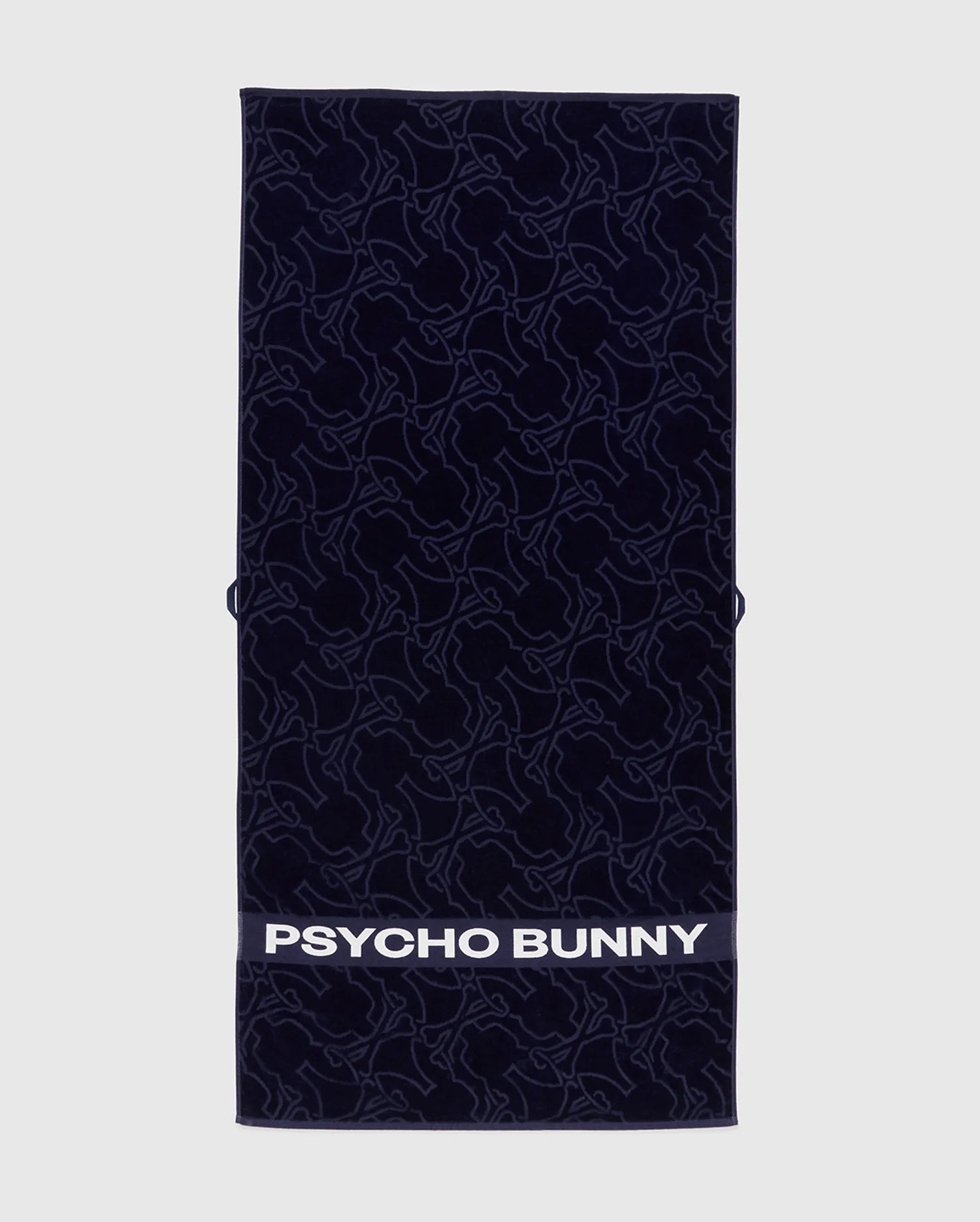The Psycho Bunny TOWEL - B6A287F200 is a navy rectangular terrycloth cotton towel featuring an abstract bunny pattern and "PSYCHO BUNNY" in bold white letters along the bottom edge.