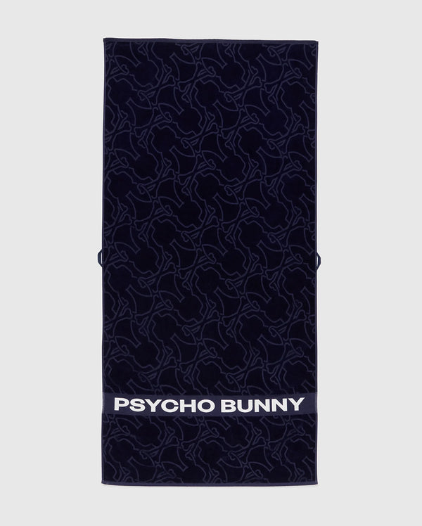The Psycho Bunny TOWEL - B6A287F200 is a navy rectangular terrycloth cotton towel featuring an abstract bunny pattern and "PSYCHO BUNNY" in bold white letters along the bottom edge.