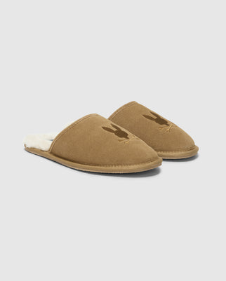 The Psycho Bunny MENS SUEDE SLIPPERS, product B6A357E200, are brown slip-ons with a genuine shearling lining for ultimate comfort. They feature an embroidered logo on the top against a plain white background, making these cozy indoor slippers a premium choice.