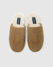 The Psycho Bunny MENS SUEDE SLIPPERS (B6A357E200) are cozy brown indoor slippers with a fluffy white shearling lining and an embroidered bunny head above crossbones. The inner sole includes a label, all displayed against a plain light gray background.