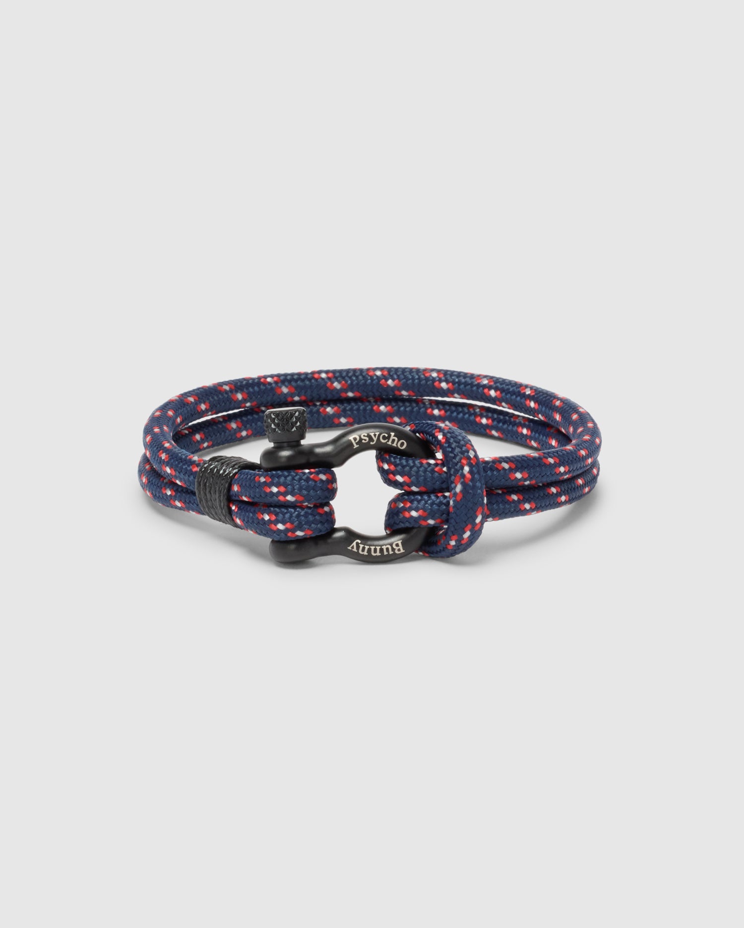 The Psycho Bunny MEN'S ROPE BRACELET - B6A368E200 is a stylish navy blue braided accessory with red and white speckles, featuring a modern lightweight design and an engraved metal clasp.