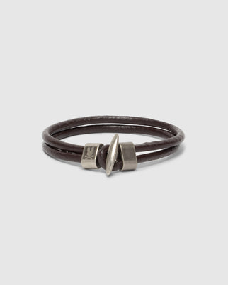 Displayed on a white background, the Psycho Bunny MEN'S LEATHER BRACELET - B6A369E200 is coiled in a circle to highlight its simple yet stylish design. Crafted from brown leather, it features a silver clasp and an engraved metal accent.