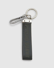 The Psycho Bunny WOVEN TAB KEYCHAIN - B6A374E200 features a silver carabiner and circular keyring. It includes a dark green, durable nylon strap with subtle matching text.