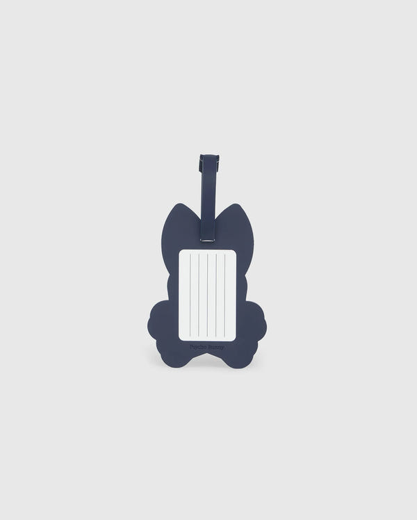 A navy blue luggage tag in the shape of a cat is pictured. The front side features a rectangular white label with blank lines for writing contact information. Made from detachable silicone, the Psycho Bunny MENS LUGGAGE TAG - B6A387B200 has a strap at the top for attaching to luggage.