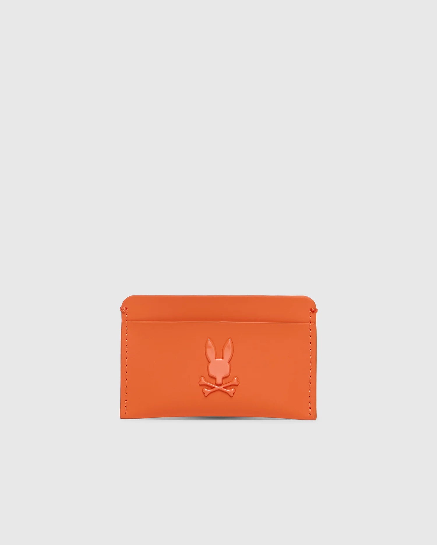 The Psycho Bunny MENS RUBBERIZED CARD CASE - B6A473F200 is a striking orange wallet featuring a debossed rabbit silhouette with crossed bones. Its minimalist design with clean stitching ensures your essentials are secure, all set against a plain white backdrop.