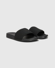 The MENS KNIT SLIDES - B6A474F200 by Psycho Bunny are black slide sandals with textured knit textile straps on a plain gray background. They feature an open-toe design, a molded footbed for comfort, and a flat, thick sole.