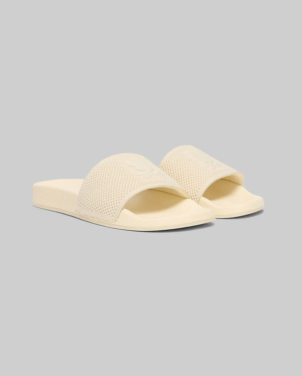 The Psycho Bunny MENS KNIT SLIDES - B6A474F200 are beige open-toe slides with textured knit straps and a molded footbed, shown on a light gray background for effortless summer comfort.