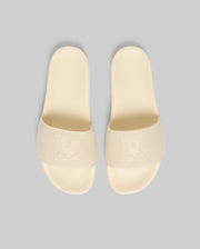 The Psycho Bunny MENS KNIT SLIDES (B6A474F200) are beige slide sandals with textured straps, an embossed bunny logo, and a molded footbed for summer comfort, shown on a light gray background.