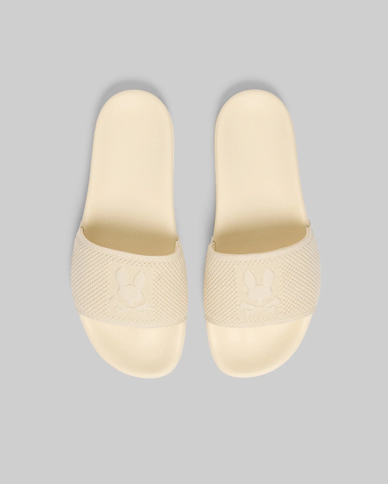 The Psycho Bunny MENS KNIT SLIDES (B6A474F200) are beige slide sandals with textured straps, an embossed bunny logo, and a molded footbed for summer comfort, shown on a light gray background.