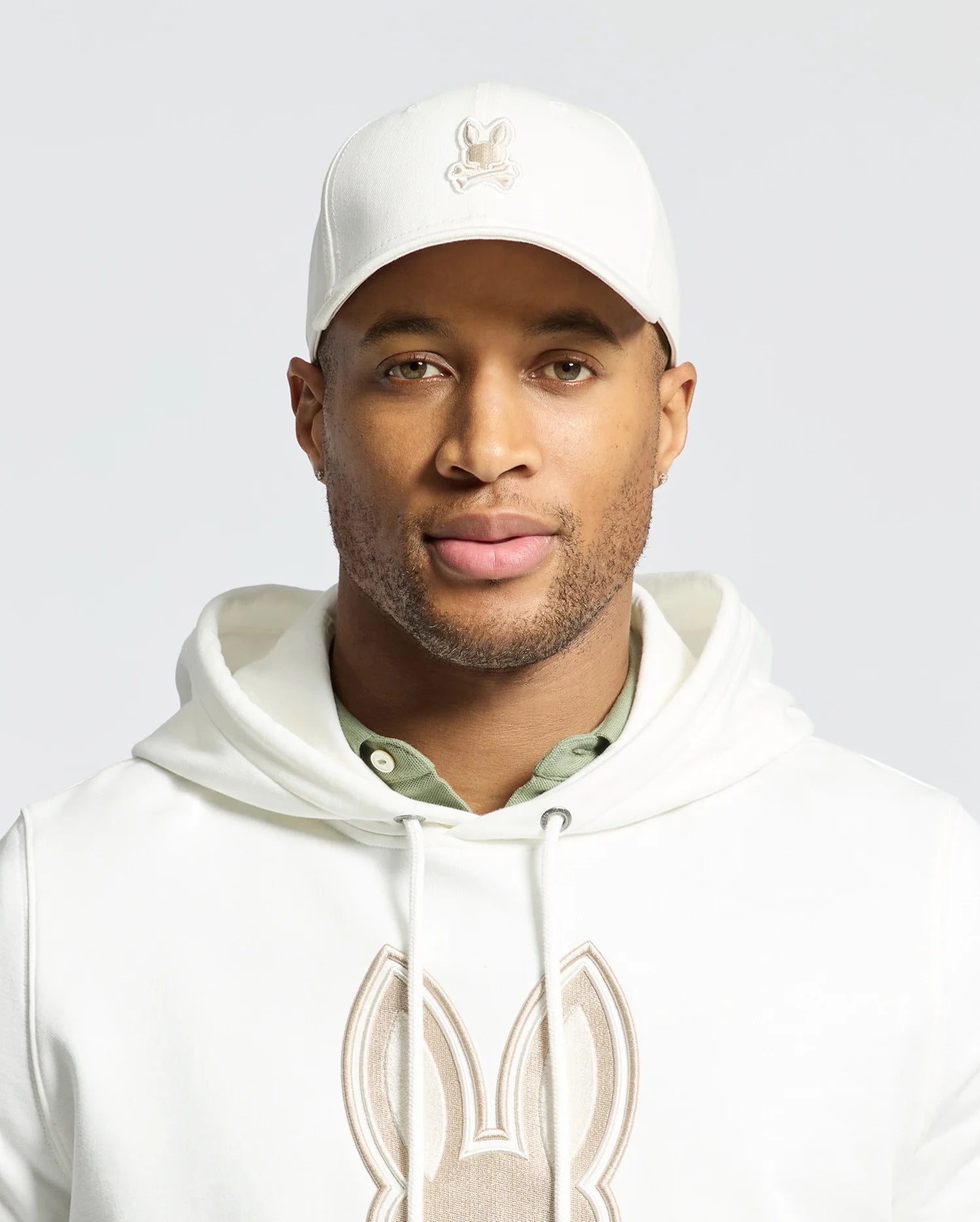 The individual sports a Psycho Bunny embroidered cap (MENS NEW CASTLE EMBROIDERED GRAPHIC BASEBALL CAP - B6A547F200), a hoodie with a rabbit design, and a green undershirt, showcasing quiet luxury through subtle details. Their expression remains neutral against a plain backdrop.