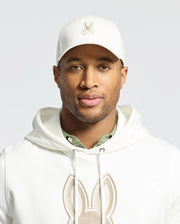 The individual sports a Psycho Bunny embroidered cap (MENS NEW CASTLE EMBROIDERED GRAPHIC BASEBALL CAP - B6A547F200), a hoodie with a rabbit design, and a green undershirt, showcasing quiet luxury through subtle details. Their expression remains neutral against a plain backdrop.