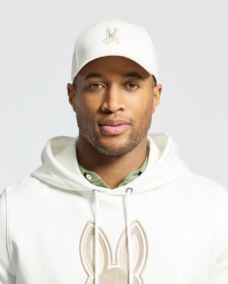 The individual sports a Psycho Bunny embroidered cap (MENS NEW CASTLE EMBROIDERED GRAPHIC BASEBALL CAP - B6A547F200), a hoodie with a rabbit design, and a green undershirt, showcasing quiet luxury through subtle details. Their expression remains neutral against a plain backdrop.