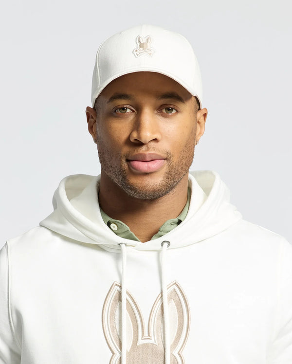 The individual sports a Psycho Bunny embroidered cap (MENS NEW CASTLE EMBROIDERED GRAPHIC BASEBALL CAP - B6A547F200), a hoodie with a rabbit design, and a green undershirt, showcasing quiet luxury through subtle details. Their expression remains neutral against a plain backdrop.