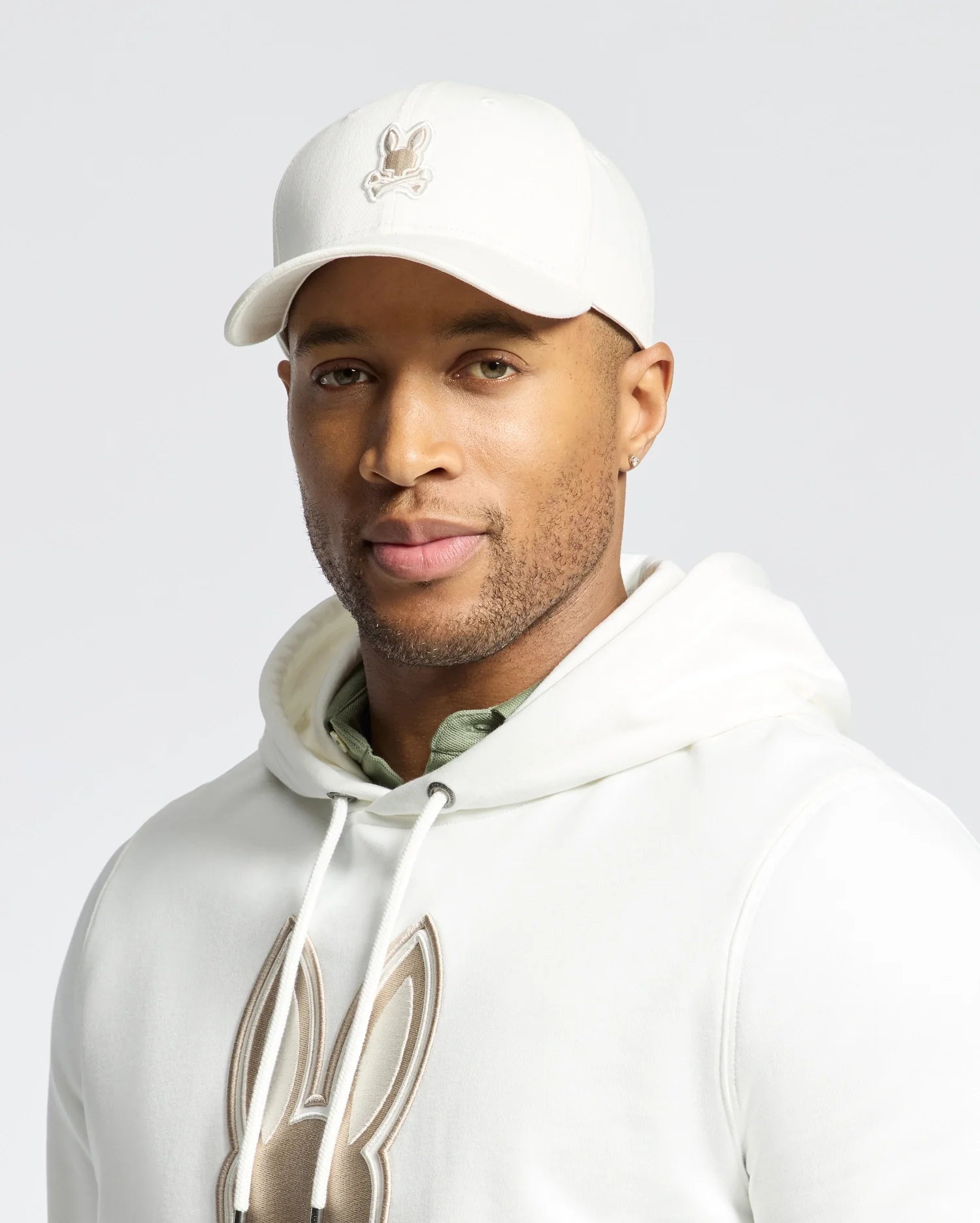 Against a plain light gray background, a person wears the Psycho Bunny MENS NEW CASTLE EMBROIDERED GRAPHIC BASEBALL CAP - B6A547F200 and a white hoodie adorned with bunny logos, exuding an air of quiet luxury.