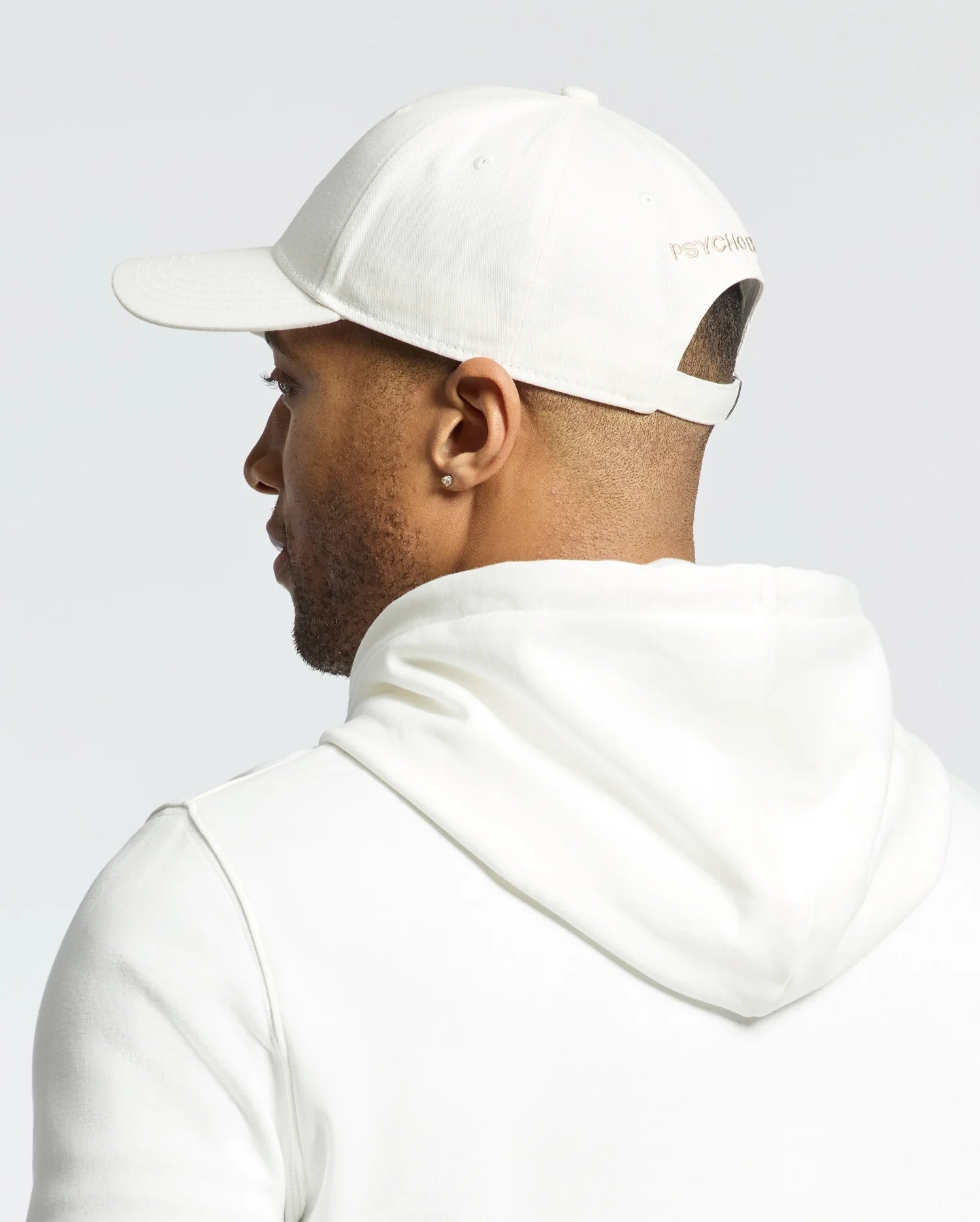 A person is wearing a white hoodie and a Psycho Bunny MENS NEW CASTLE EMBROIDERED GRAPHIC BASEBALL CAP - B6A547F200, with small text on the back. The scene is captured from behind against a plain, light-colored background.
