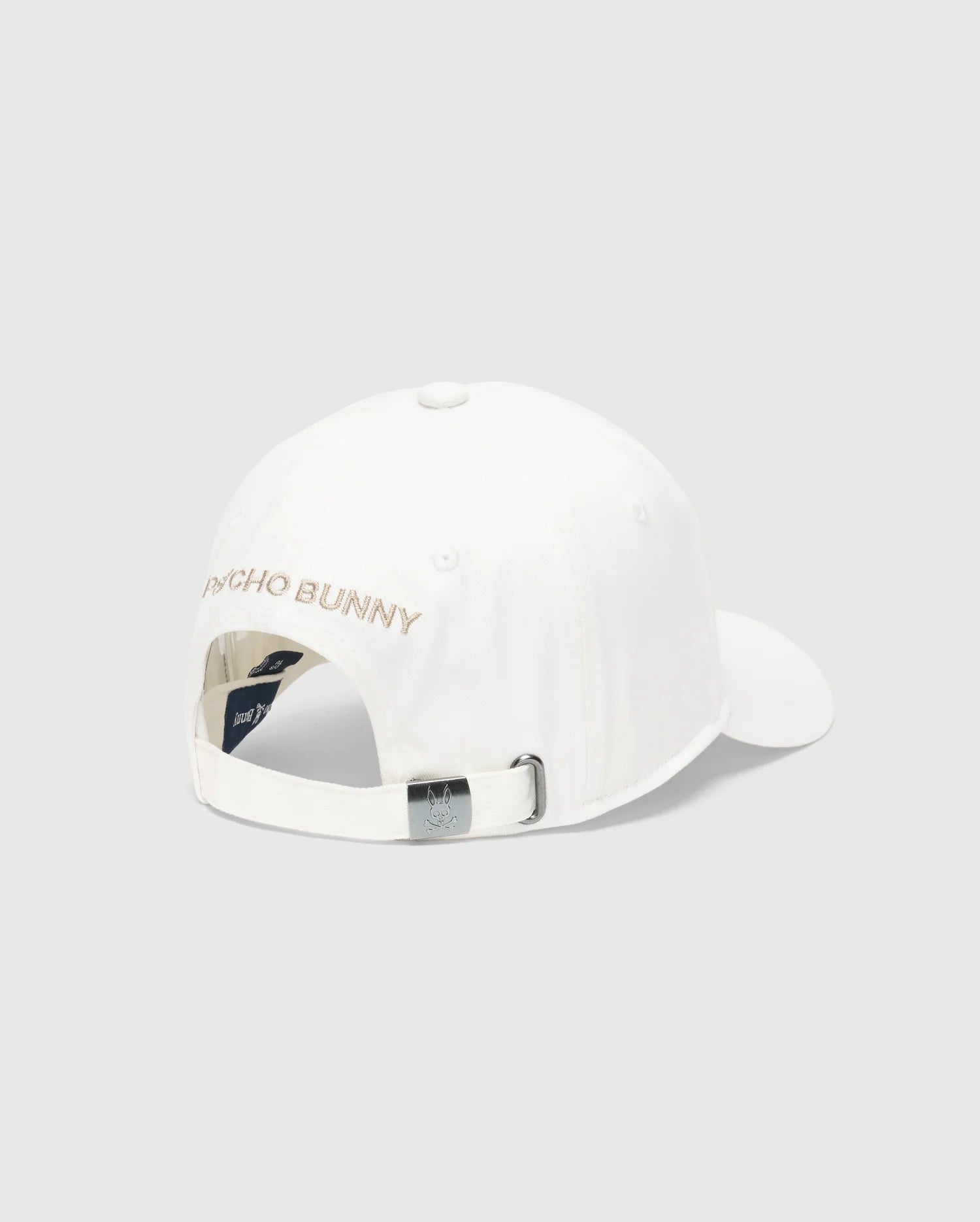 The MENS NEW CASTLE EMBROIDERED GRAPHIC BASEBALL CAP by Psycho Bunny features a back view of an embroidered cotton twill design with "PSYCHO BUNNY" in light tan, embodying quiet luxury. It includes an adjustable strap with a metal buckle, a top button, and ventilation eyelets.