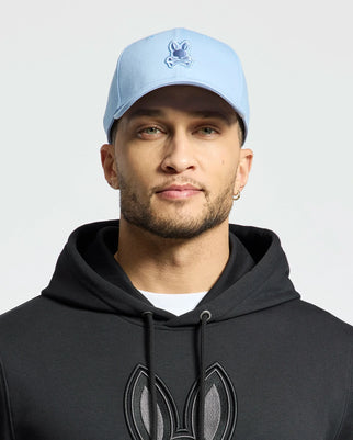 A bearded man wears a Psycho Bunny New Castle embroidered cap in light blue cotton twill and pairs it with a black hoodie featuring the same bunny design. He accessorizes with a small hoop earring against a plain white background, embodying quiet luxury.