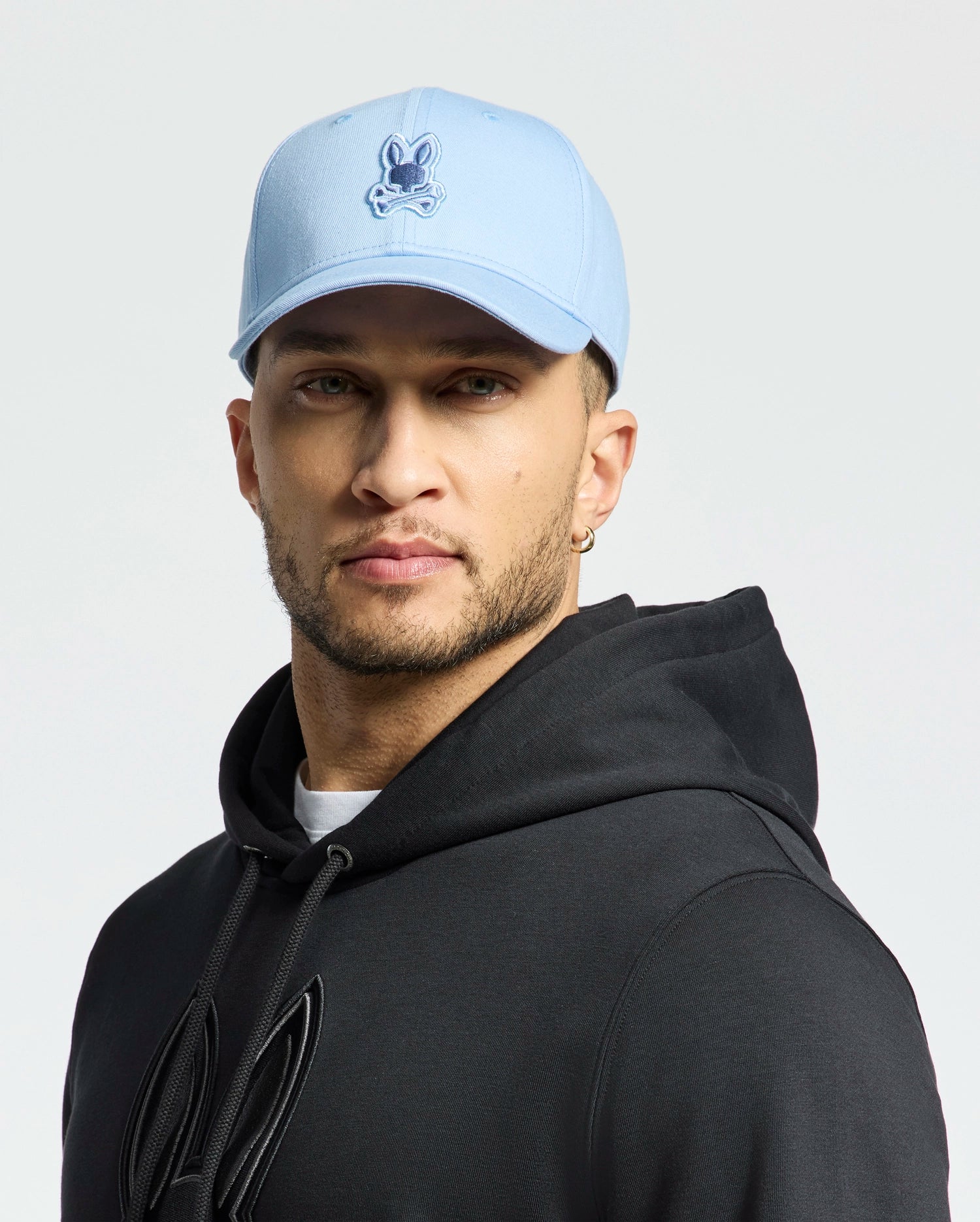 A person wearing a Psycho Bunny MENS NEW CASTLE EMBROIDERED GRAPHIC BASEBALL CAP in light blue, featuring a cartoon logo, faces the camera. They have short facial hair, wear a black hoodie, and stand against a plain white backdrop, embodying understated quiet luxury.